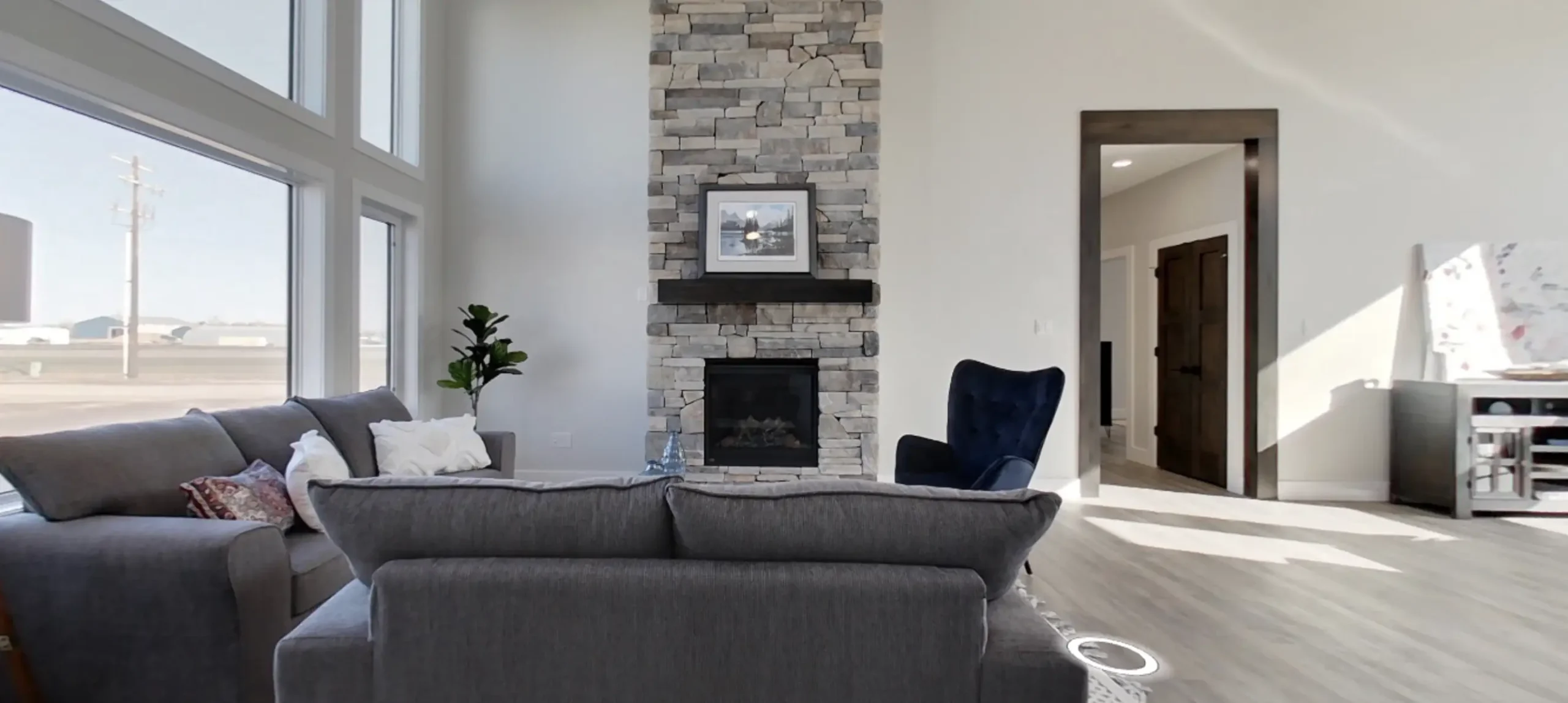 RTM home living room in Alberta or Saskatchewan with floor to ceiling stone fireplace, large grid windows, gray sectional sofa, and a blue accent chair.